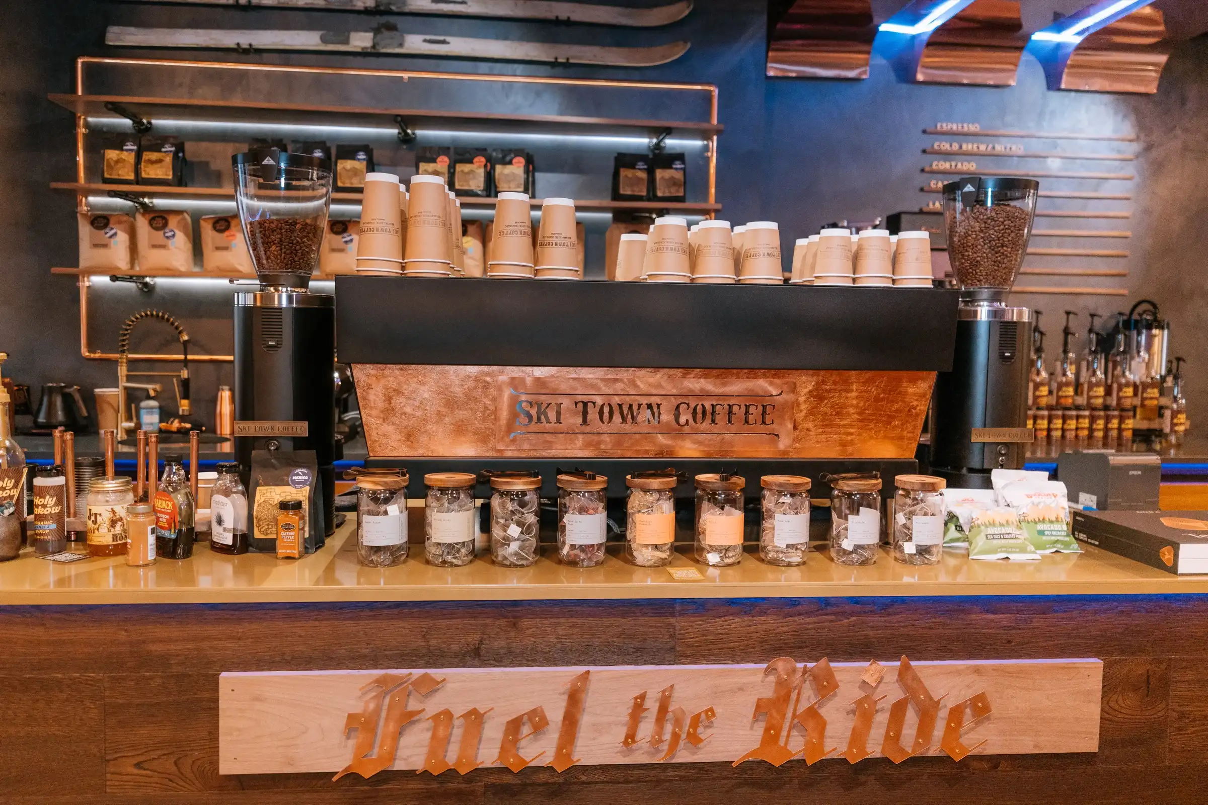 The bar at Ski Town Coffee: a copper-fronted espresso machine, shelves of coffee, and a carved Fuel the Ride sign on the bar front.