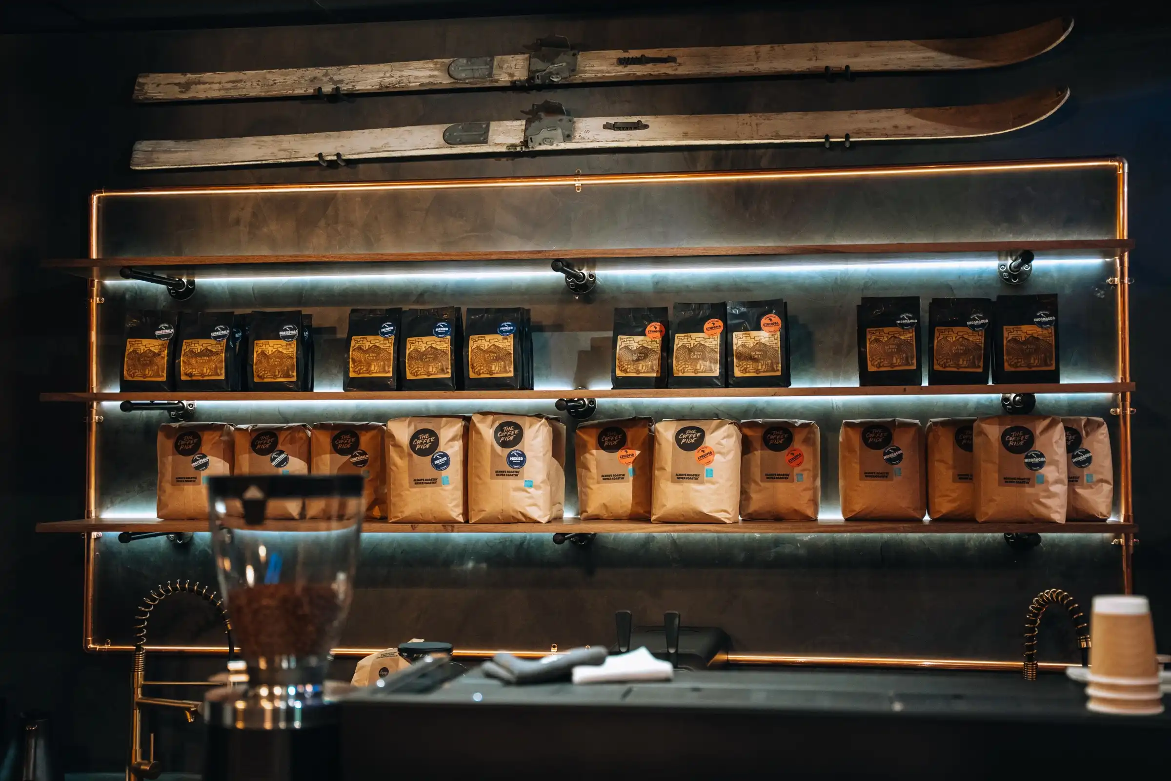 Bags of coffee on copper pipe shelves lit with blue light, with two old wooden skis mounted above.