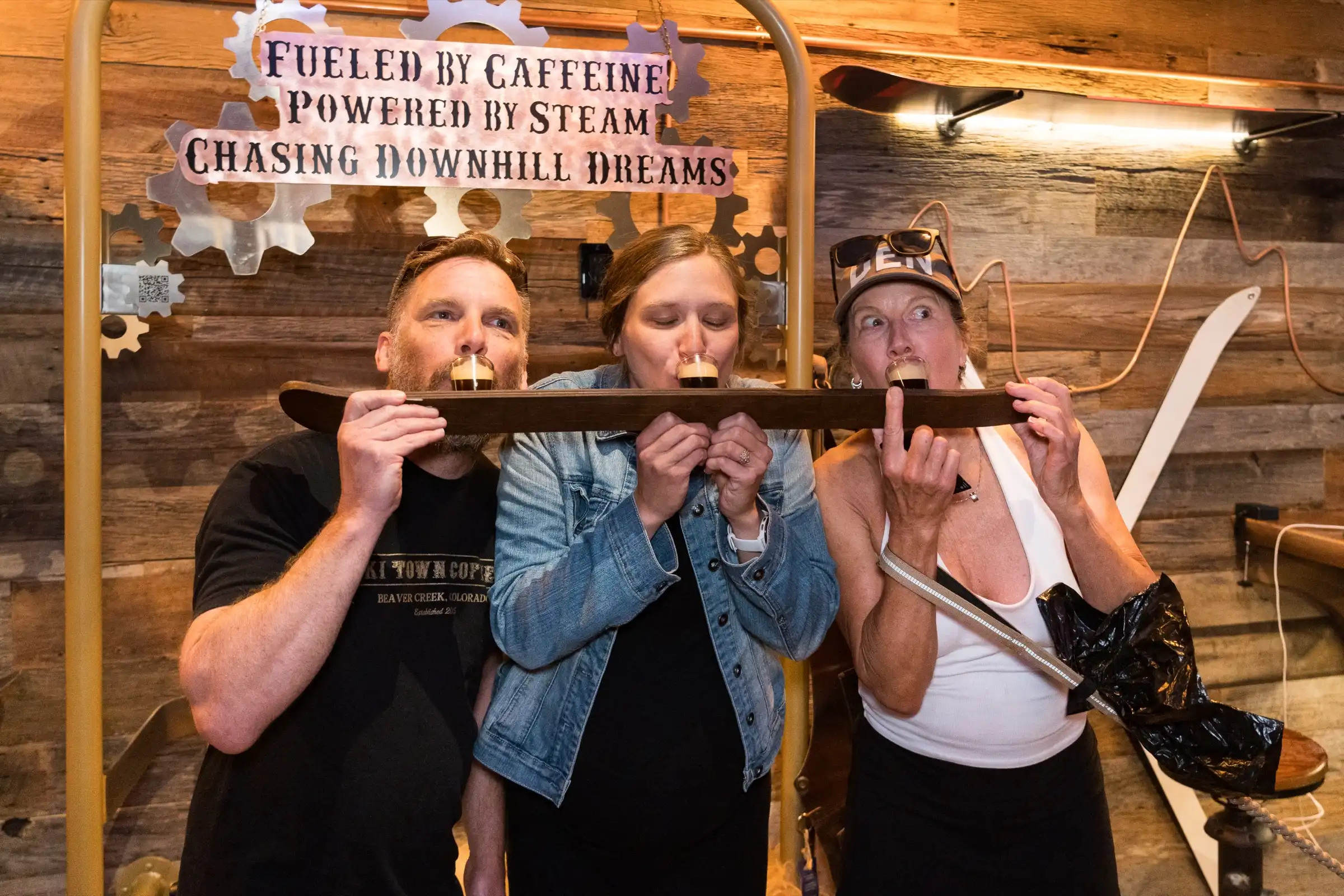 Three people drinking from a shotski, a ski with shot glasses mounted on it, in front of our Fueled by caffeine, powered by steam, chasing downhill dreams sign.